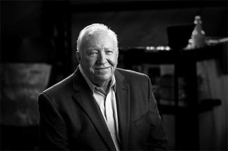 A grayscale photo of Jim Harris, Sr. smiling. Behind him is a shelf full of cleaning supplies.