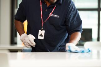 Janitronics cleaning staff wiping down a table. One hand is holding a spray bottle, and the other is wiping the table with a microfiber cloth.