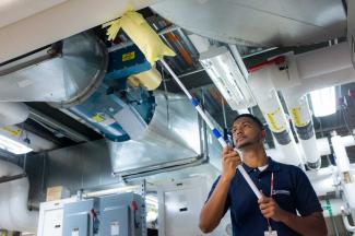 A Janitronics employee dusting around ductwork in an industrial environment.