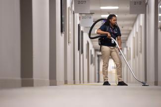 a Janitronics janitor vacuuming a carpeted hallway using a backpack vacuum and sweeping the nozzle over the carpet