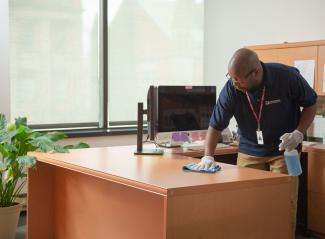 A Janitronics custodian wiping down a desk in an office with a microfiber cloth and cleaning spray