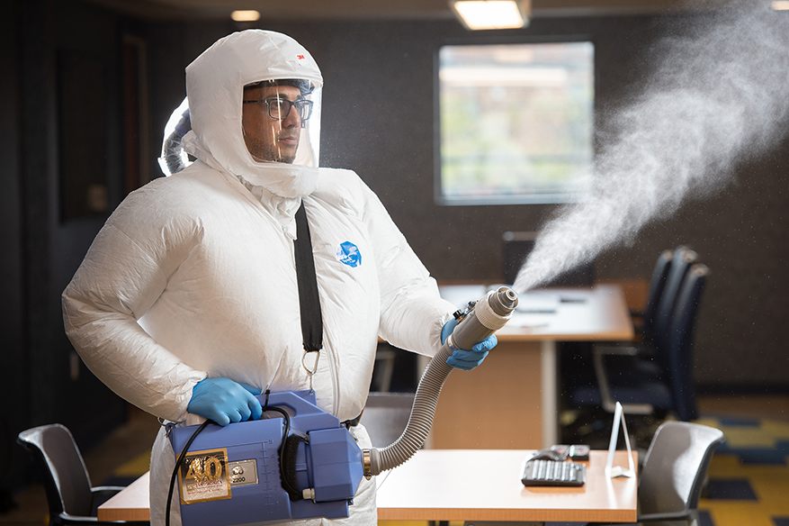 a Janitronics cleaning technician in a hazmat-like suit, using electrostatic spray to sanitize a room